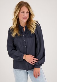 Marinblauwe blouse met bloemenkantdetails bij de halslijn, lange pofmouwen en knoopsluiting. Soepele, lichte stof.