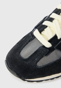 Fekete velúr edzőcipő, amely hálós panelekből, krém színű fűzőkből és texturált talpból áll. A dizájn finoman kidolgozott varrási részleteket és lekerekített orrformát tartalmaz.