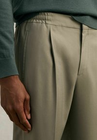 Pantalons plissés pour hommes en tissu vert olive, dotés d'une taille élastique relaxée et de poches latérales, montrés avec une main reposant sur la cuisse.