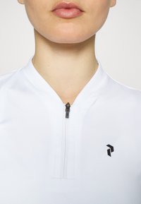 Haut de sport blanc avec un col haut, doté d'une courte fermeture éclair et d'un petit logo noir sur le côté gauche. Texture lisse, design minimal.
