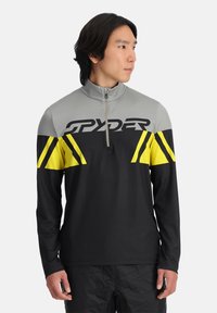 Zwart langemouwen top met grijze kraag en gele accenten. Heeft een kwart rits en het "SPYDER"-logo over de borst in dikke letters.