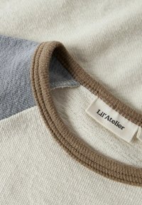 Baumwollhemd mit einem Design in Beige und hellgrau in Blockfarben. Es hat einen braunen gerippten Kragen und ein Etikett mit der Aufschrift "Lil'Atelier".