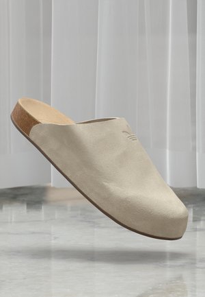 Μπεζ σουέτ slip-on κλογ με φελλό σόλα, αιωρούμενο πάνω από ένα γυαλιστερό μάρμαρο πάτωμα και λευκές διαφανείς κουρτίνες στο φόντο.