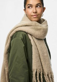 Sciarpa di lana beige con una consistenza morbida e bordi frangiati, appoggiata su un indumento esterno verde. Il design della sciarpa è semplice e accogliente.