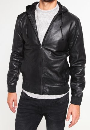 Man die een zwarte leren zip-up hoodie draagt over een lichtgrijze gebreide trui en zwarte spijkerbroek, staand tegen een effen achtergrond.