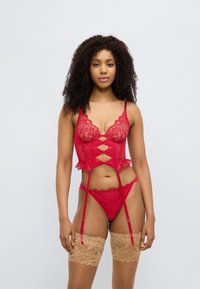 Σετ κόκκινου δαντελένιου εσωρούχου με περίπλοκα λουλουδάτα σχέδια, ρυθμιζόμενους ιμάντες και λεπτομέρειες ζαρτιέρας, συνδυασμένο με μπεζ δαντελένιες κάλτσες μέχρι τους μηρούς.