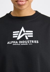 Juodas medvilnės džemperis su baltu grafiniu logotipu, turinčiu tris juosteles ir tekstą "ALPHA INDUSTRIES" bei "KNOXVILLE, TENNESSEE, U.S.A."