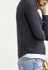 Ciemnoniebieski sweter dzianinowy o fakturowanym wzorze, okrągłym dekolcie, ściągaczu u dołu i długich rękawach. Zestawiony z jasnoniebieskimi dżinsami.