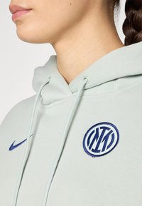 Lysegrønn hettegenser laget av myk stoff, med en blå Nike-logo og et brodert sirkulært emblem med et blått og hvitt design.