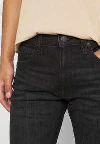 Jeans en denim noir avec une coupe ajustée, présentant une fermeture à boutons et des poches latérales. Portés avec un t-shirt beige.