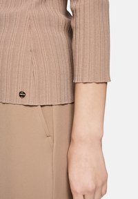 Nahaufnahme eines beigen, gerippten Strickpulloverärmels und einer passenden Hosentasche mit einem kleinen runden Metallmarkenanhänger nahe dem Saum.