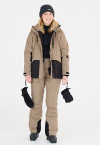 Vaaleanbeige laskettelutakki mustilla yksityiskohdilla, vetoketjulliset taskut ja säädettävä huppu, yhdistettynä yhdistelemiseen vaaleanbeigeihin laskettelu-housuihin ja mustiin lapasiin.