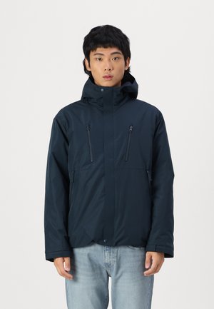 Veste imperméable navy avec un col haut, une capuche ajustable et deux poches poitrine zippées. Texture lisse et design minimaliste.