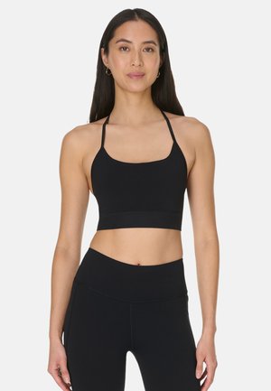 Vrouw draagt een zwarte halter-sportbeha en zwarte leggings met hoge taille, staand rechtop tegen een effen lichte achtergrond.
