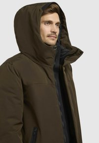 Brun isoleret jakke med hætte, som har lynlås foran, trykknapper og sorte detaljer. Glat tekstur og tætsiddende design.