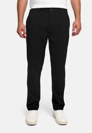 Mann trägt eng anliegende schwarze Hose und weiße Sneaker, steht vor einem schlichten hellen Hintergrund.
