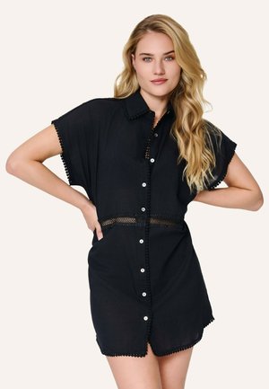 Donna bionda che indossa un vestito corto nero con bottoni e dettagli in pizzo in vita, in piedi con le mani sui fianchi davanti a uno sfondo neutro.