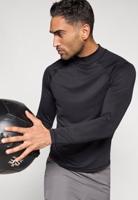 Μαύρο αθλητικό μπλουζάκι με μακρύ μανίκι και ψηλό γιακά, εφαρμοστή γραμμή και ομαλή υφή. Το άτομο κρατάει μια μαύρη γυμναστική μπάλα με υφή.