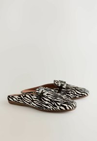 Schwarz-weiße Zebra-Muster-Hausschuhe mit glatter Textur und metallischen Schnallenakzenten an der Oberseite. Weiches, flaches Sohlen-Design.