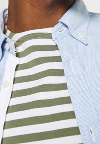 Lichtblauw geruit overhemd met knopen, gedragen over een groen-wit gestreept T-shirt. Voorzien van een kraag, knoopsluitingen en zachte texturen.