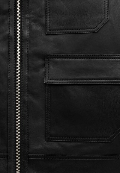 Close-up of black leather jacket showing silver zipper and two stitched pockets with flaps on smooth textured surface.