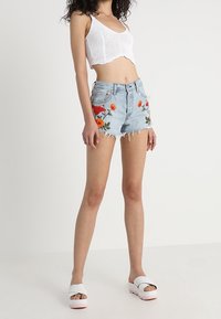 Džínové šortky s květinovou výšivkou v živých barvách, roztřepený lem, doplněné bílým pleteným krátkým topem a bílými sandály na platformě.