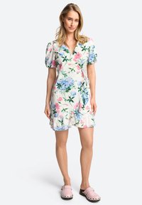 Bloemen wikkeljurk van lichte stof met roze en blauwe bloemen en groene bladeren. Korte pofmouwen en een gerimpelde zoom. Draagt met roze klompen.