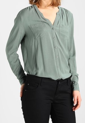 Femme portant un chemisier vert clair à boutons avec deux poches poitrine et un pantalon noir, debout devant un fond blanc uni.