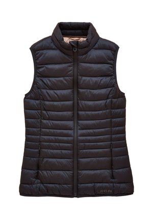 Kvinde stående med krydsede ben iført en lyserød sweater med et blomsterbil-design, blå jeans, navy vest og beige støvler.