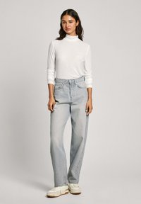 Un col roulé blanc à côtes, un jean taille haute bleu clair avec jambes larges, et des baskets blanches avec des accents beiges, sur un fond neutre.