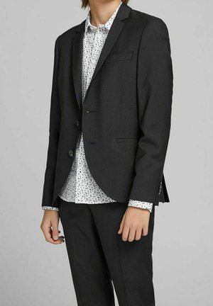 Person iført en sort skræddersyet blazer over en hvid skjorte med knapper og sort plettet mønster, kombineret med sorte bukser.