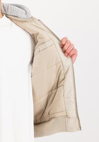 Beige zippered jacket with quilted lining, grey hood, elastic cuffs, and smooth outer material. Lightly padded for insulation.