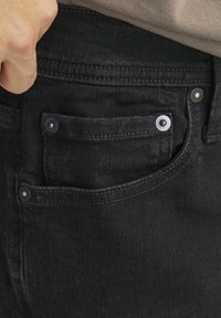 Black denim jeans featuring a front pocket with silver hardware, contrast stitching, and a textured fabric. Close-up view of the pocket area.