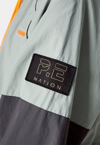 Chaqueta con bloques de color en gris claro y gris oscuro, que presenta un parche texturizado con letras doradas. Incluye un cierre de cremallera en un brillante color naranja.