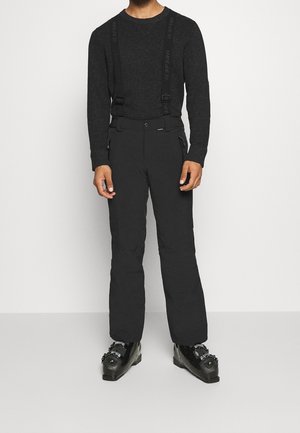 Mężczyzna ubrany w czarne szelki, czarne ocieplane spodnie, czarny dziany sweter oraz buty narciarskie, stojący na białym tle.