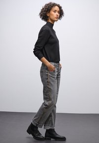 Haut noir à col roulé côtelé, jeans en denim gris avec surpiqûres dorées sur le côté, ourlet retroussé et bottines noires, sur fond neutre.