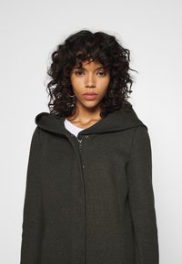 Manteau à capuche vert foncé en tissu texturé, doté d'une fermeture éclair à l'avant, au design minimaliste avec des bords arrondis.