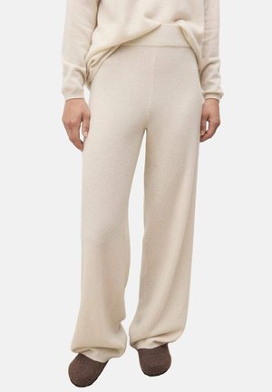 Donna che indossa pantaloni in maglia beige larghi abbinati a un maglione coordinato e pantofole marroni, in piedi contro uno sfondo bianco.