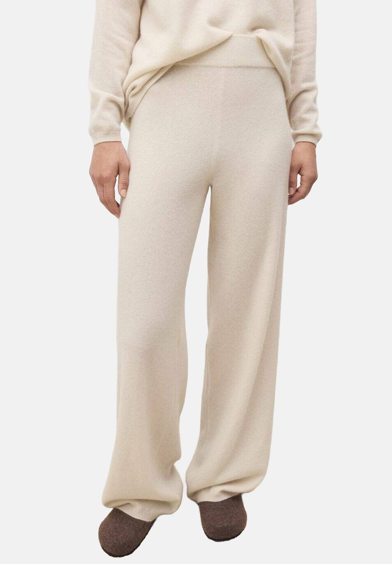 Donna che indossa pantaloni in maglia beige larghi abbinati a un maglione coordinato e pantofole marroni, in piedi contro uno sfondo bianco.