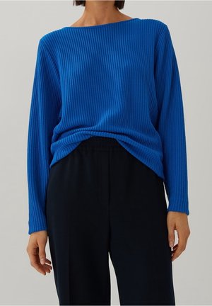 Person iført en lyseblå ribbet bluse med lange ærmer, stoppet ned i løse sorte bukser, stående mod en ensfarvet lysegrå baggrund.