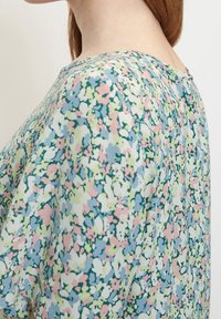 Detailní pohled na osobu v blůze s pastelovým květinovým vzorem v modré, zelené, růžové a bílé barvě na jednobarevném pozadí.