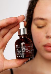 Botella de vidrio ámbar oscuro con un tapón gotero plateado, etiquetada como "Ampoule Intensiva de Reparación de Caracol" en texto blanco, sostenida por una mano.