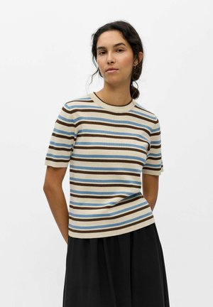 Femme portant un haut en maille crème à manches courtes avec des rayures horizontales bleues et brunes, assorti à une jupe noire, debout devant un fond blanc uni.