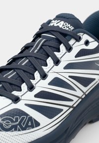 Bílo-modré běžecké boty s texturovaným svrškem, síťovinovými panely, reflexními akcenty a polstrovanou podrážkou s viditelným šněrovacím systémem.