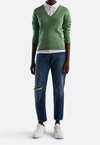 Grön v-ringad tröja, vit skjorta med krage under, i kombination med slitna blå jeans och vita sneakers. Avslappnad outfit med lager av texturer.