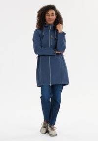 Laivansinis vandenį atstumiantis džemperis su užtrauktuku priekyje, gaubtu, raištelių akcentais ir dviem šoninėmis kišenėmis, derinamas su mėlynais džinsais ir smėlio spalvos batais.