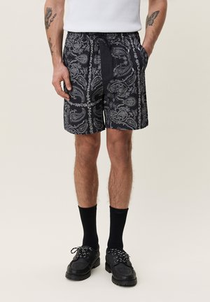 Man die zwarte shorts met paisley-patroon draagt, zwarte sokken, zwarte schoenen en een wit mouwloos hemd, staand met handen in de zakken.