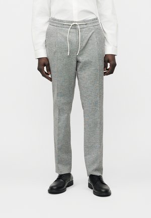 Mand iført lysegrå bukser med snøre, hvid skjorte med lange ærmer og sorte pæne sko mod en ensfarvet baggrund.