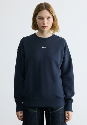 Młoda kobieta o jasnobrązowych włosach, ubrana w granatową bluzę z logo "BOSS", stojąca na jednolitym jasnoszarym tle.