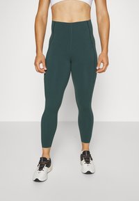 Aukštos liemens tamsiai žalios legginsai, pasižymintys lygiu, elastingu medžiagos paviršiumi, su šoninėmis detalėmis, derinami su baltais sportiniais batais.